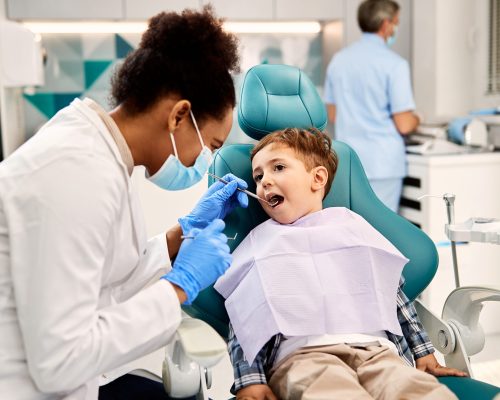 Black female dentist examining small boy's teeth during dental procedure at dentist's office. Focus is on boy.